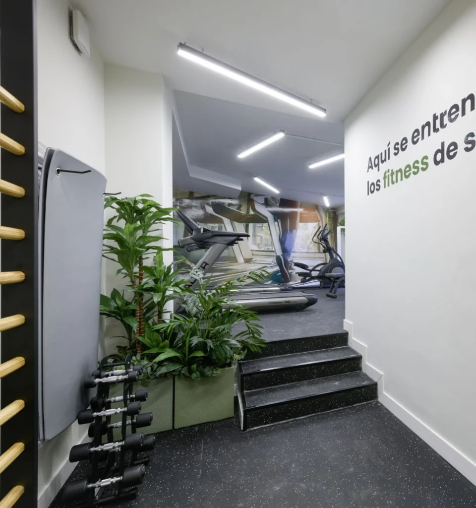 Interior de un gimnasio moderno con cintas de correr, bicicletas estáticas, mancuernas, una escalera de madera para ponerse en forma, plantas verdes y texto motivador en español en la pared. El espacio está bien iluminado, con paredes blancas y suelo negro.