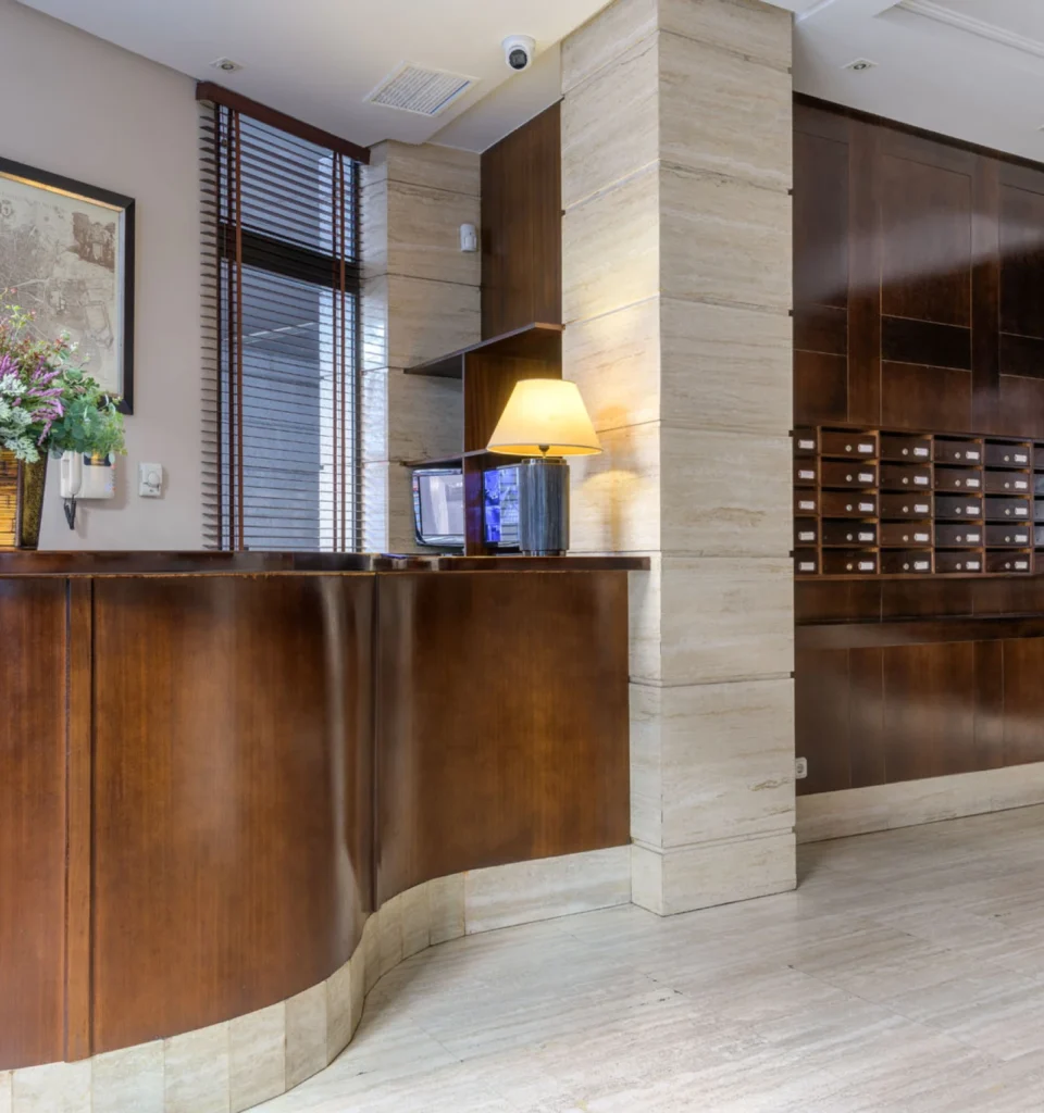 Recepción de hotel con un mostrador curvado de madera, una lámpara, buzones en la pared, un jarrón con flores y suelo y paredes de baldosas de color neutro. A la izquierda hay una ventana con persianas de madera.