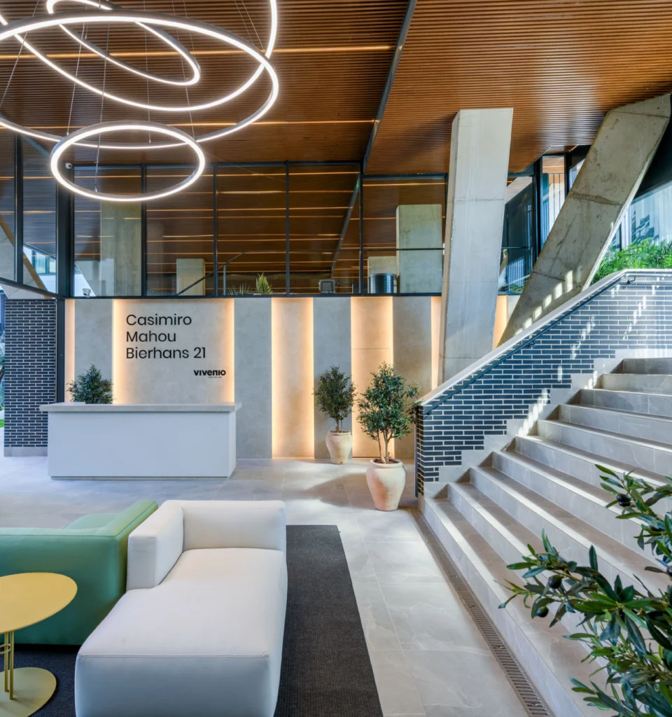 Vestíbulo de oficina moderno con un sofá blanco, mesas verdes y amarillas, plantas en macetas, lámparas circulares iluminadas en el techo, una escalera con azulejos azules y un cartel que reza Casimiro Mahou Bierhaus 21 en la pared detrás de la recepción.