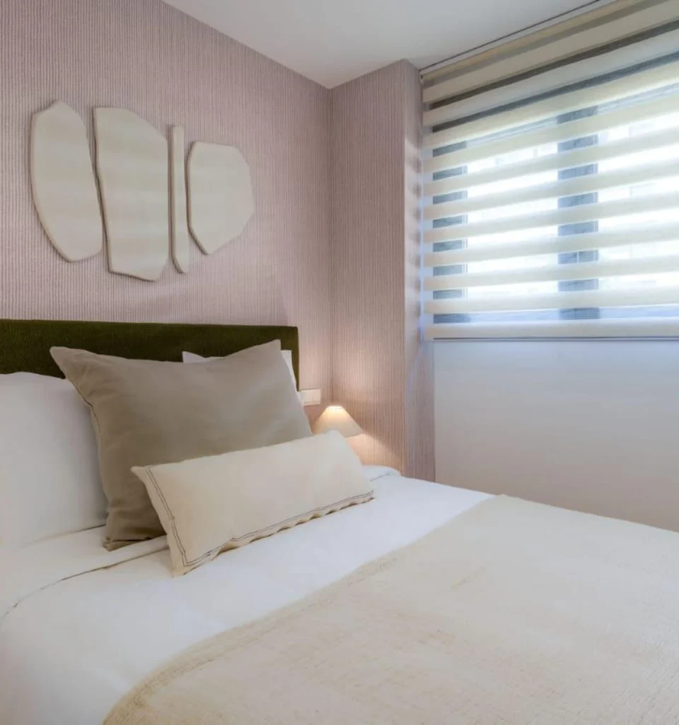 Un dormitorio con una cama individual, almohadas y mantas de colores neutros, decoración en la pared sobre el cabecero, una pequeña lámpara de mesilla y una ventana con persianas horizontales que deja pasar la luz natural.