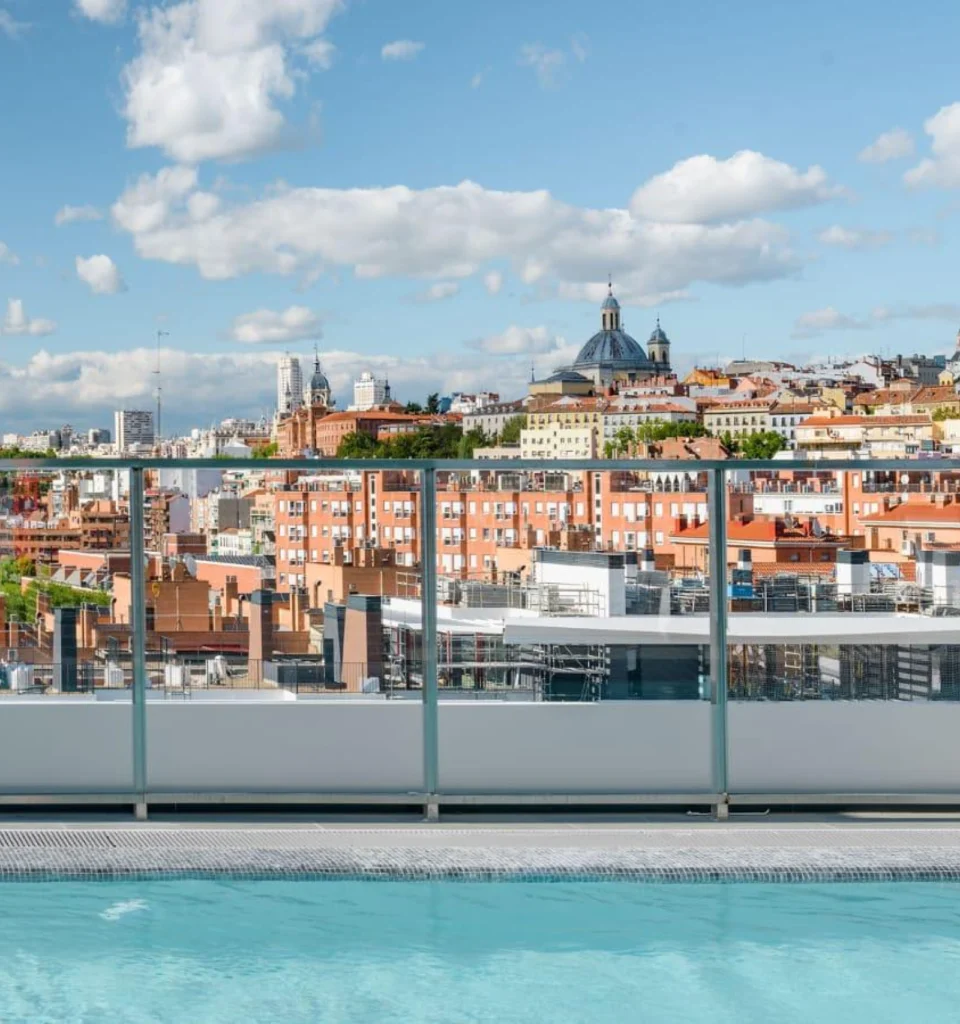 Una piscina en la azotea con barandilla de cristal domina un paisaje urbano de edificios y cúpulas de tejados rojos bajo un cielo azul con nubes dispersas.