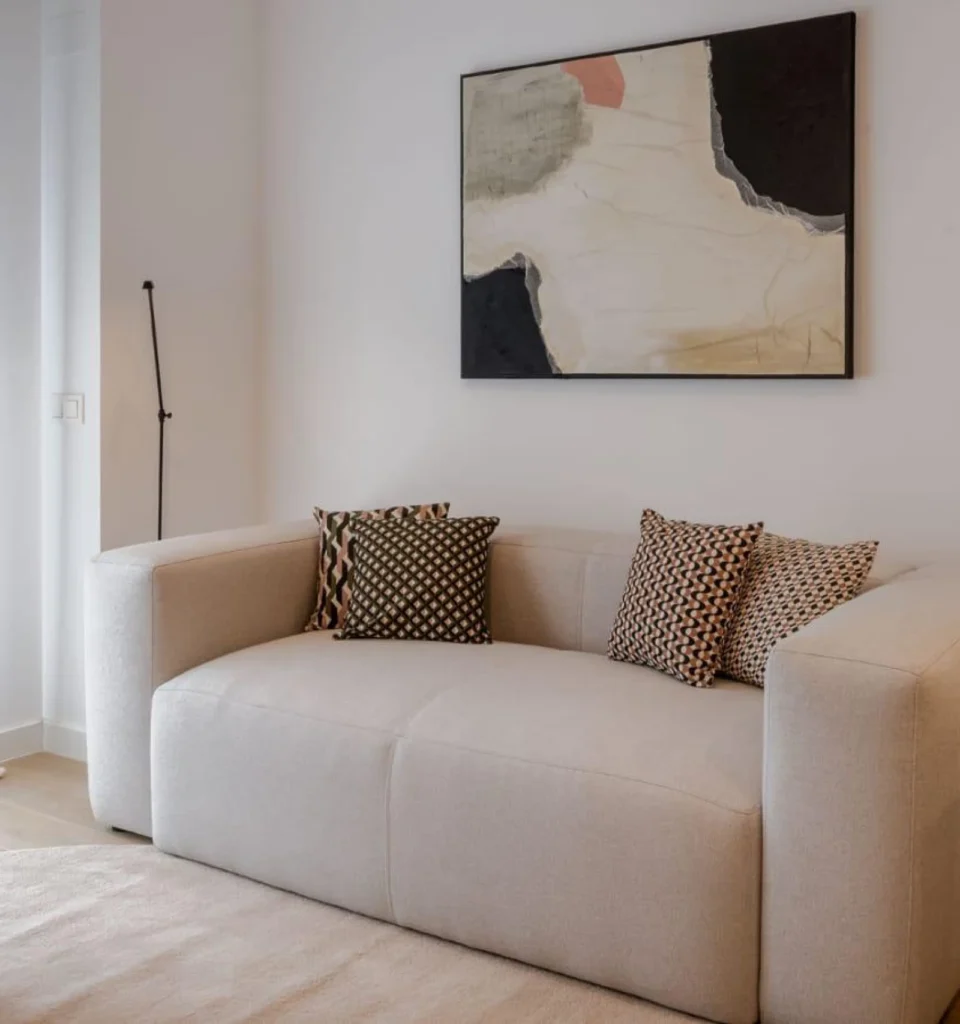 Un salón con un sofá beige claro, cuatro cojines estampados, arte abstracto en la pared y una lámpara de pie en la esquina.