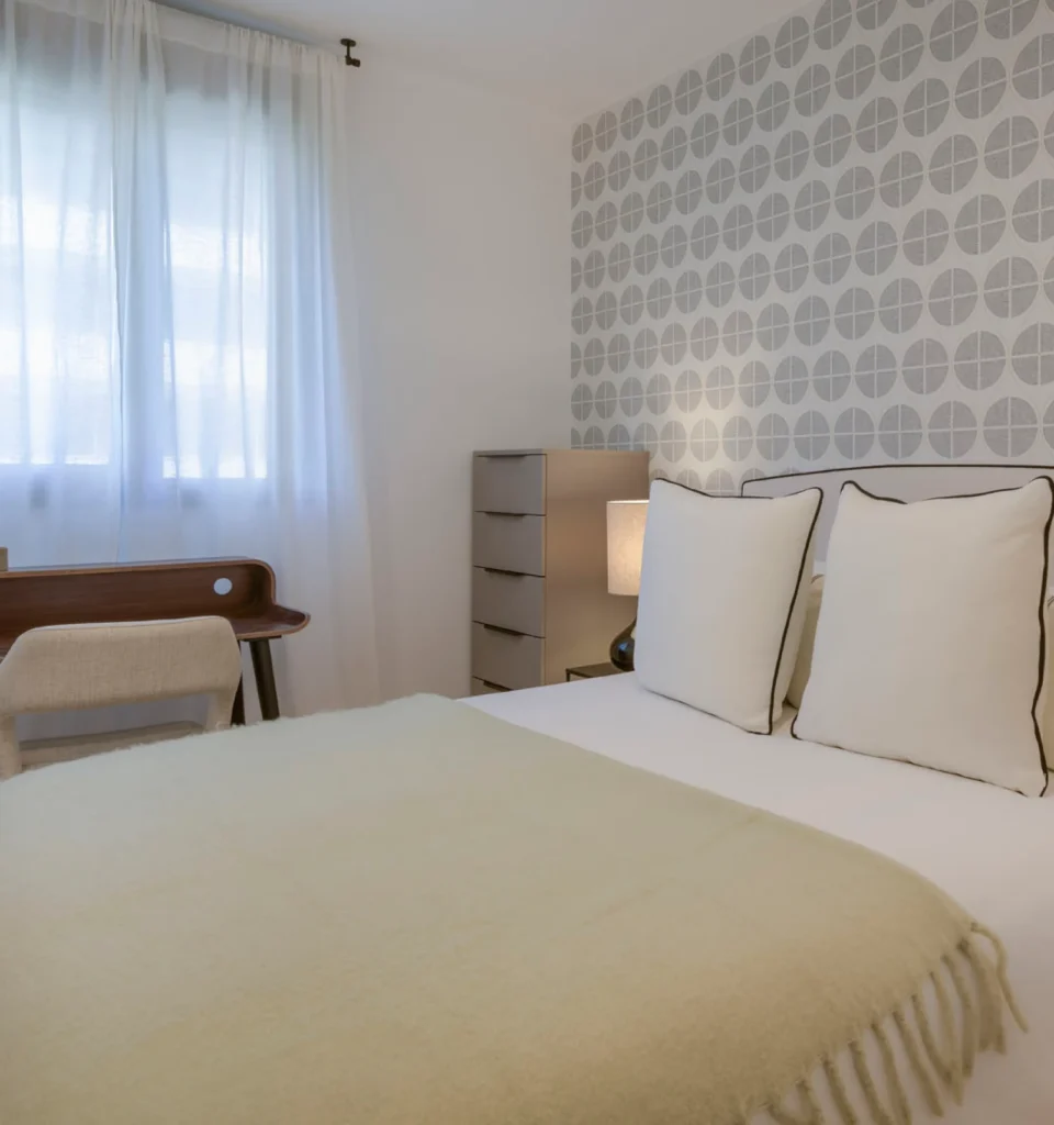 Un dormitorio con una cama individual, dos almohadas grandes, una manta de color beige claro, una pared estampada, una cómoda pequeña, un escritorio de madera y una silla acolchada cerca de una ventana con cortinas transparentes.