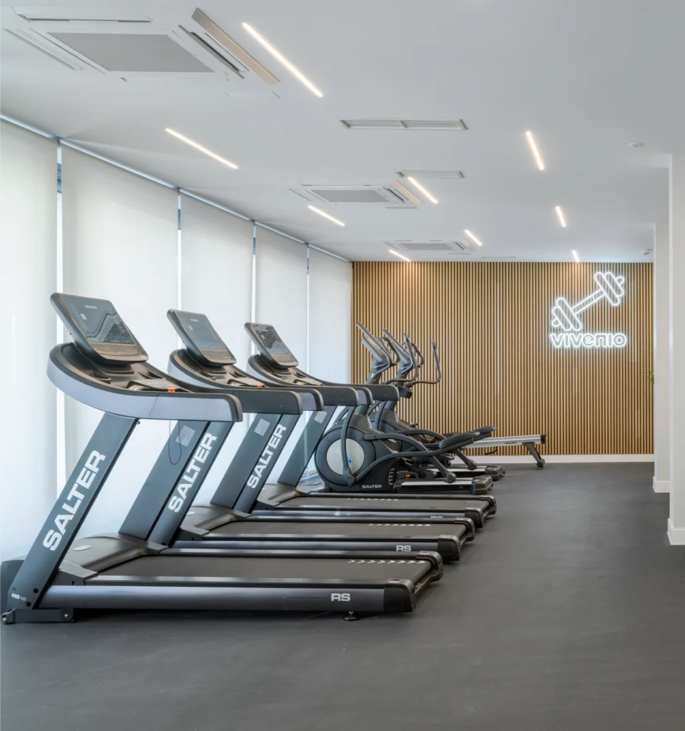 Un gimnasio moderno con cuatro cintas de correr y dos máquinas elípticas frente a grandes ventanales. La sala tiene una pared de acento de paneles de madera con un letrero de neón de Viverlo y una iluminación brillante y uniforme.