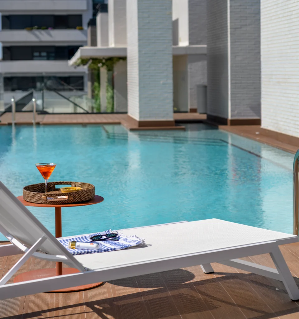 Una piscina cuadrada, una tumbona blanca junto a una pequeña mesa redonda que sostiene un cóctel y una bandeja de mimbre.