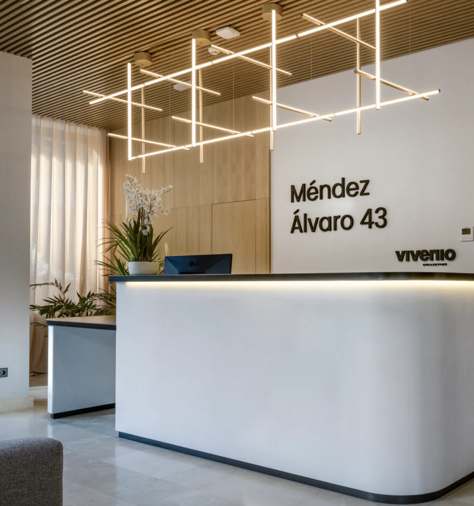Moderna zona de recepción con un escritorio curvo blanco, un monitor de ordenador negro, plantas en macetas y lámparas decorativas en el techo. En la pared aparece el rótulo "Méndez Álvaro 43" y "Viventiio" en letras negras.