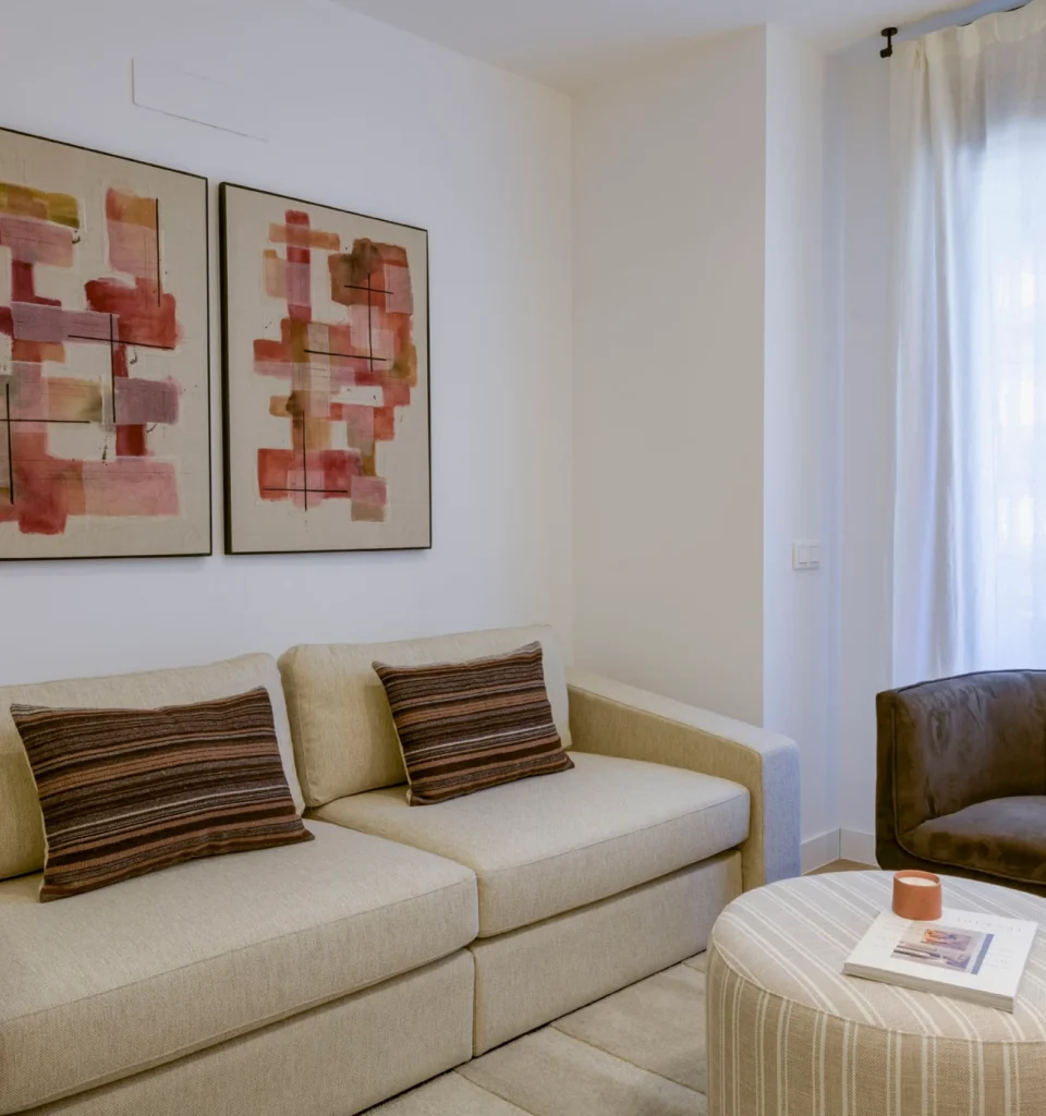 Un salón moderno con un sofá beige, dos cojines a rayas, arte abstracto en la pared, una mesa redonda con un libro y una vela, un sillón oscuro y cortinas blancas largas.