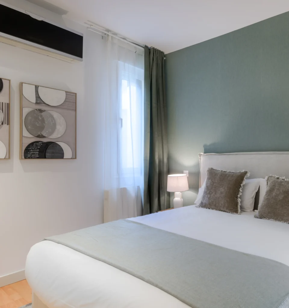 Un dormitorio con una cama individual, una pared verde y otra blanca, dos almohadas grises, una mesilla de noche con una lámpara, cortinas blancas transparentes, obras de arte en la pared y un aparato de aire acondicionado.