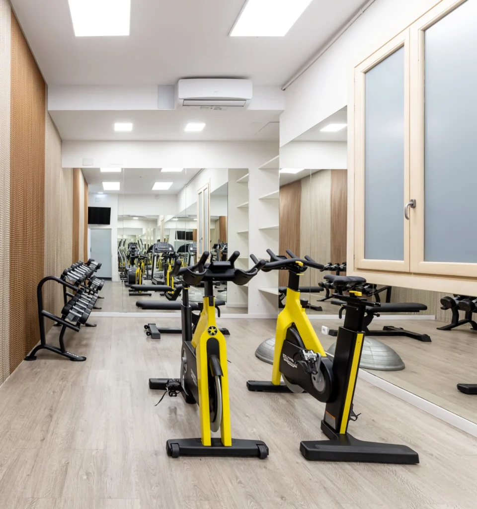 Una moderna sala de gimnasio con dos bicicletas estáticas amarillas, un estante con mancuernas, un gran espejo de pared, suelo de madera clara y paredes neutras con estanterías y ventanas.
