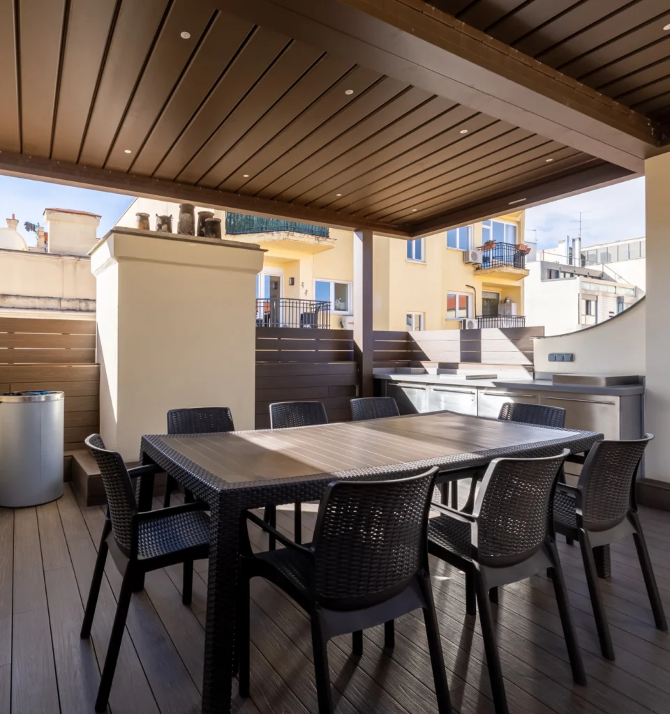 Un patio exterior con una pérgola de madera, una mesa de comedor rectangular, ocho sillas negras, suelo de madera y una parrilla empotrada, rodeado de edificios y escalones que conducen a una cubierta superior.