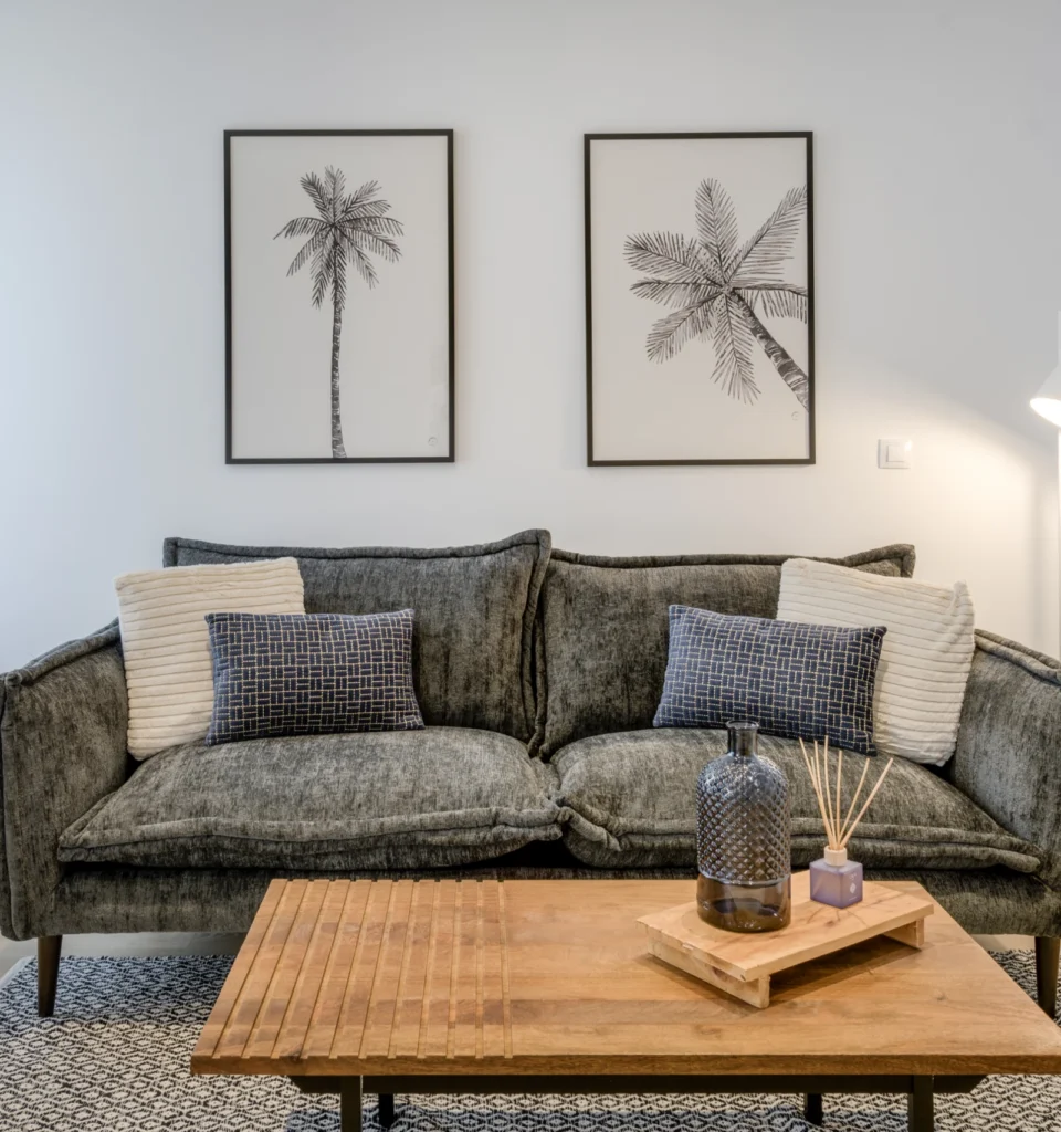 Una acogedora sala de estar con un sofá de terciopelo gris, cojines estampados y una mesa de centro de madera con artículos de decoración. Dos cuadros decorativos en la pared.