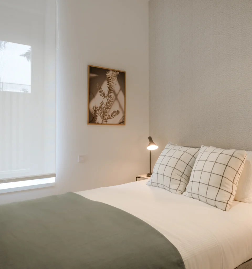 Dormitorio con una cama individual con sábanas blancas, una manta verde y dos cojines a cuadros. Sobre la mesilla de noche hay una lámpara y un cuadro. Ventana con cortina transparente.