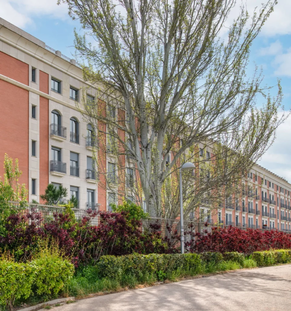 Un gran edificio de apartamentos de varias plantas con paredes rojas y beige se alza detrás de un árbol alto y una variedad de arbustos de hojas verdes y rojas, bajo un cielo parcialmente nublado.