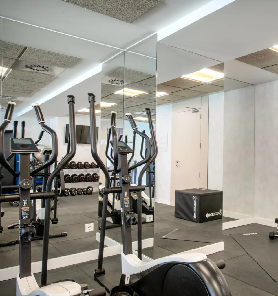 Un gimnasio moderno con bicicletas estáticas, una pared de espejos, soportes para mancuernas, un televisor, una caja pliométrica y suelo de caucho, todo ello en un espacio luminoso y limpio con paredes blancas y azulejos en el techo.