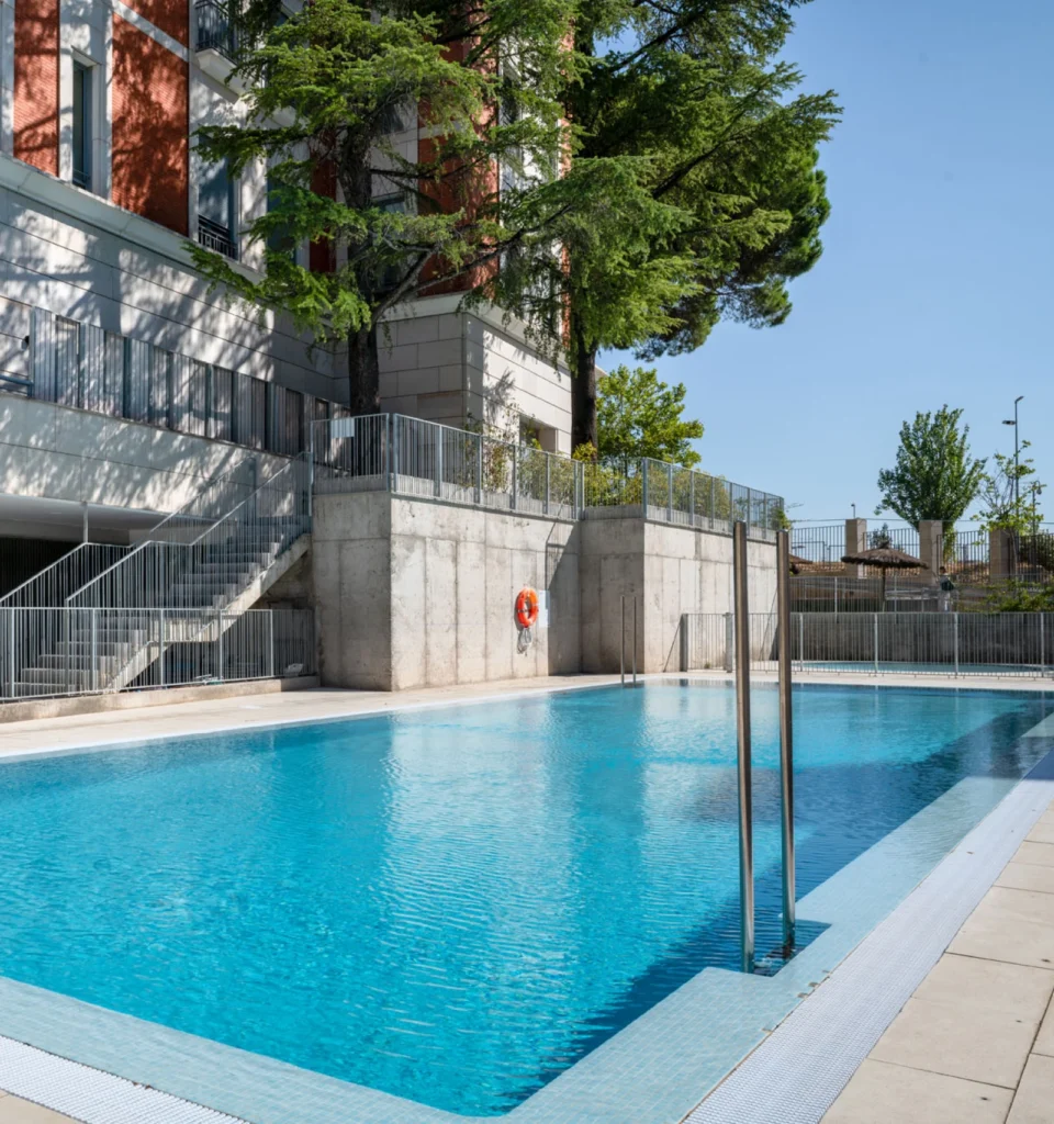 Una piscina al aire libre, limpia y rectangular, de agua azul clara, junto a un edificio moderno, rodeada de barandillas metálicas, muros de hormigón y árboles, bajo un cielo despejado. Se ven escalones y un salvavidas.