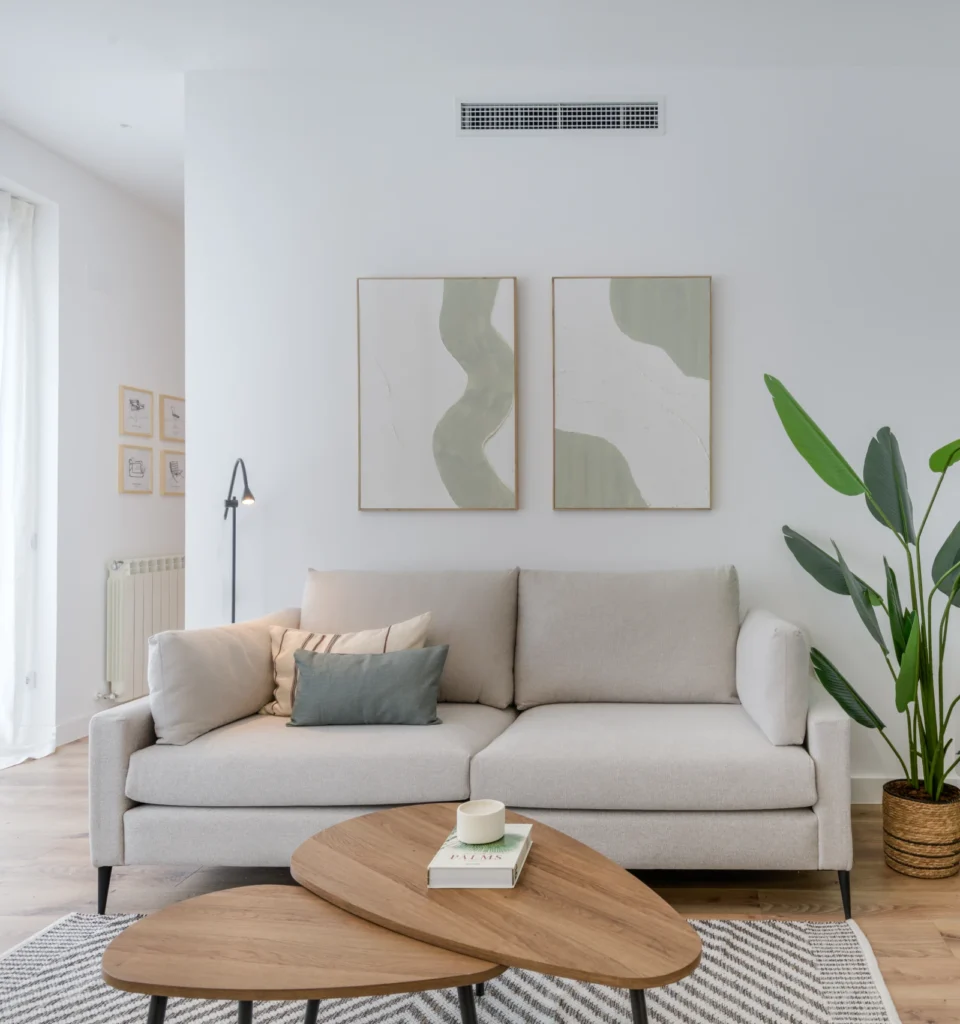 Un salón con un sofá beige, cojines decorativos, dos cuadros abstractos en la pared, una planta verde y una mesa de centro de madera con libros y una vela.