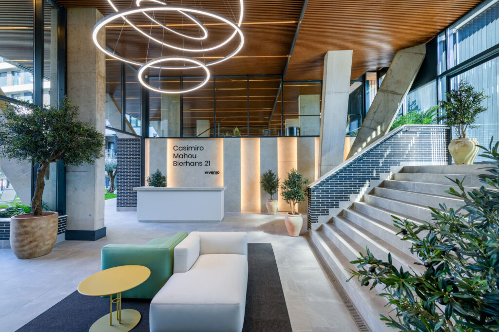 Vestíbulo de oficina moderno con mobiliario contemporáneo, un mostrador de recepción blanco, lámparas LED circulares, plantas en macetas, escaleras y una pared en la que se expone Casimiro Mahou Bierhans 21. Grandes ventanales dejan entrar la luz natural.