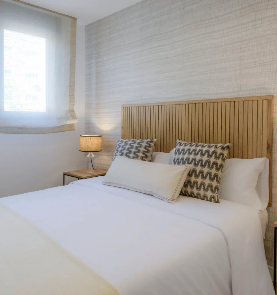 Dormitorio con una cama individual con sábanas blancas y cojines estampadas y de color beige. Cabecero de madera, una mesilla de noche con lámpara y una ventana con cortinas transparentes.
