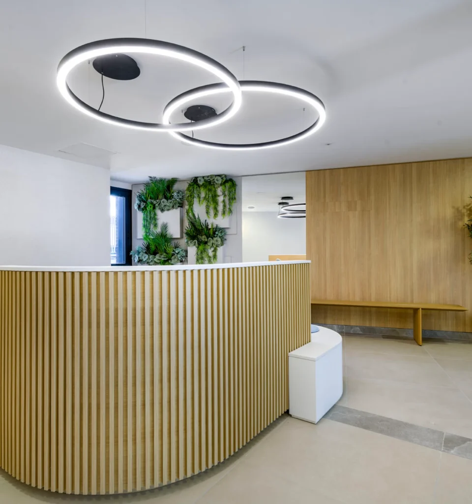 Moderna zona de recepción con un mostrador de madera curvado, luces LED circulares en el techo, plantas verdes montadas en la pared, un banco de madera y suelos y paredes de colores neutros.