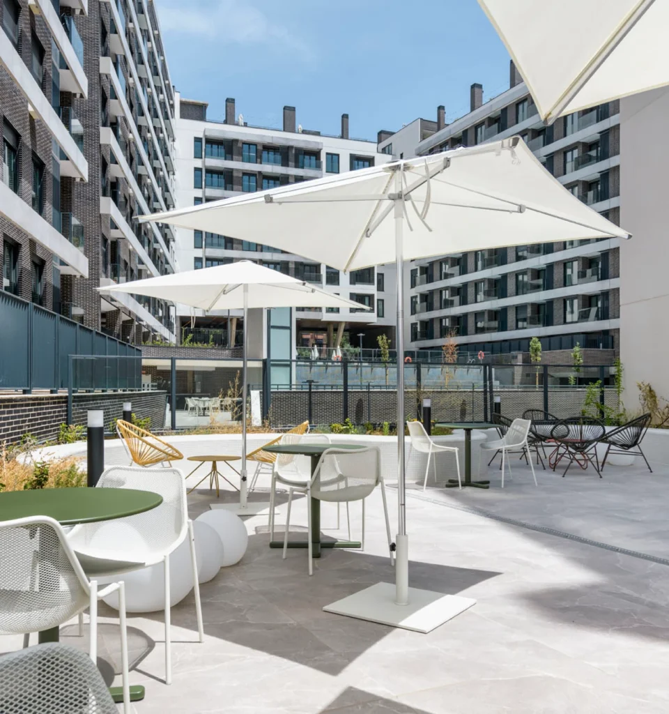 Patio de una urbanización residencial al aire libre con sombrillas blancas, mesas y sillas, rodeada de altos edificios de apartamentos.