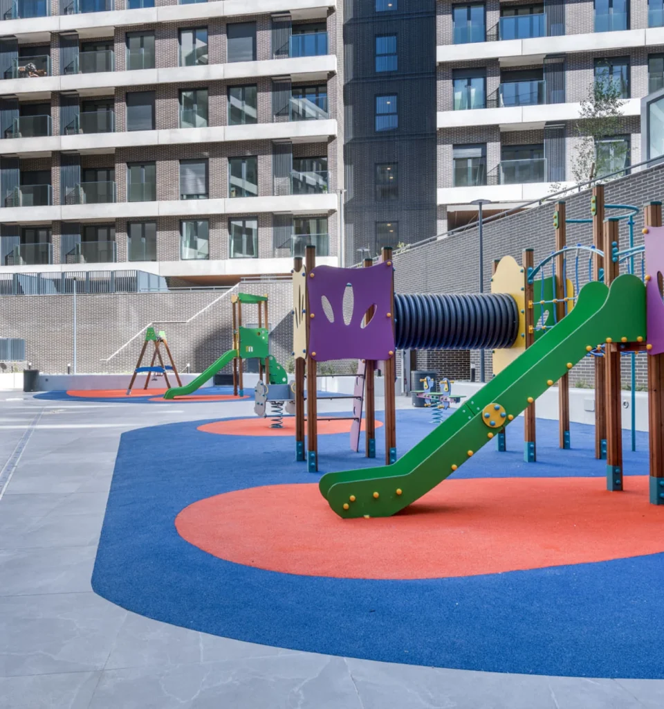 Un parque infantil en el interior de una urbanización residencial al aire libre con coloridos toboganes y columpios, sobre suelo de caucho azul y naranja.