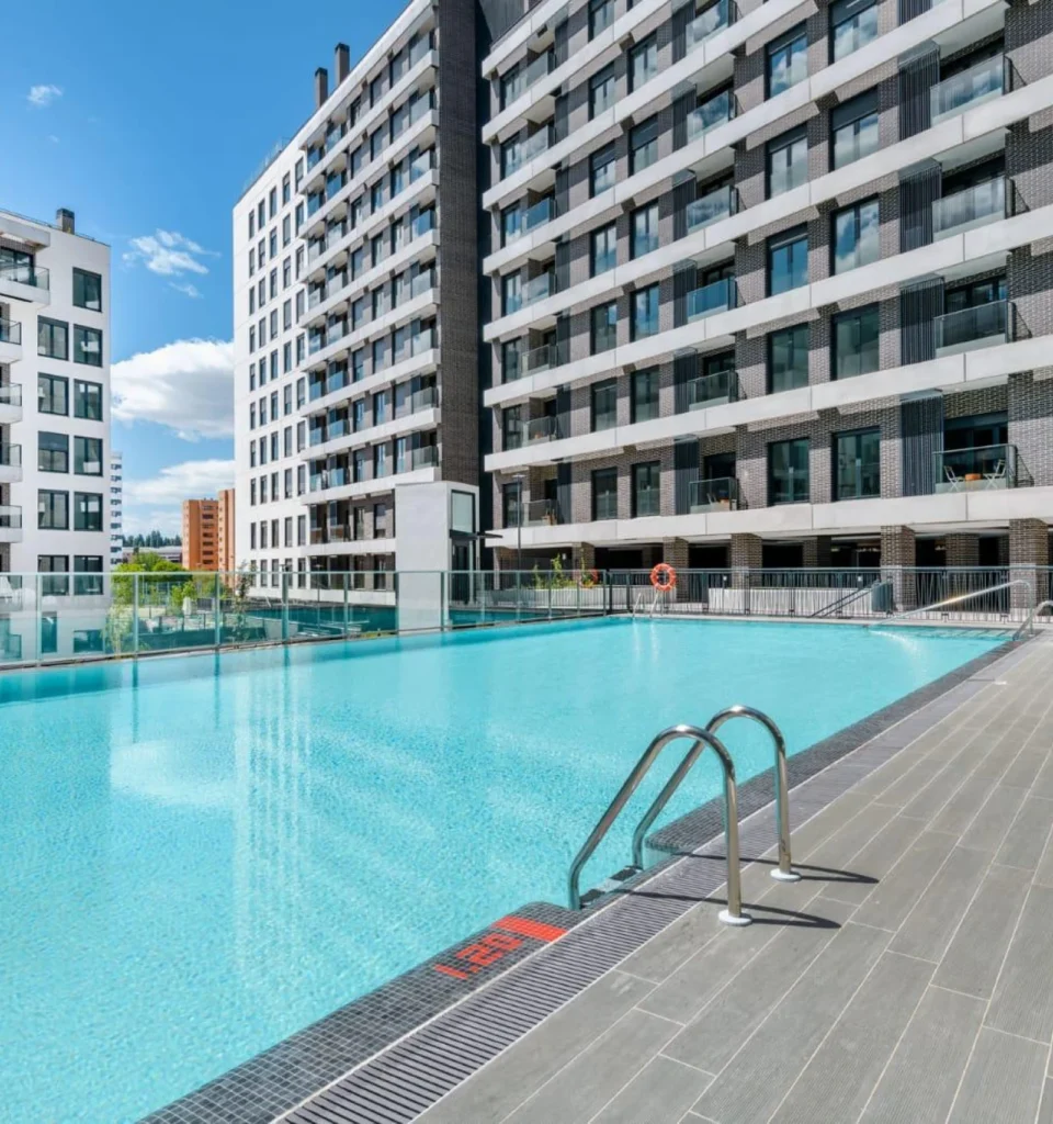 Piscina al aire libre con escalera metálica, agua azul clara y modernos edificios de apartamentos al fondo.