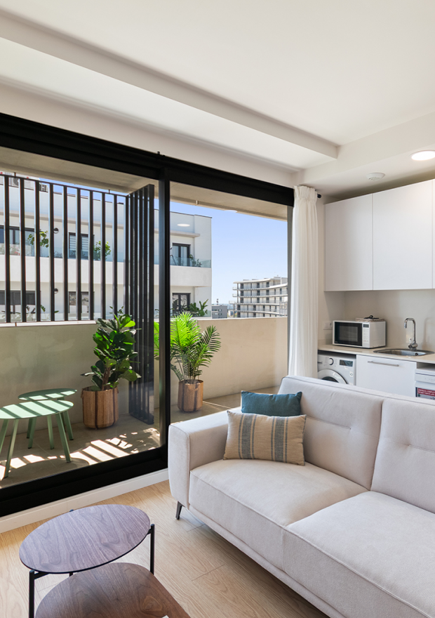 Sala de estar de un apartamento moderno con un sofá claro, una pequeña cocina y un balcón con macetas, sillas verdes y una mesa. Las grandes puertas correderas de cristal dejan entrar la luz natural y muestran otros edificios de apartamentos del exterior.