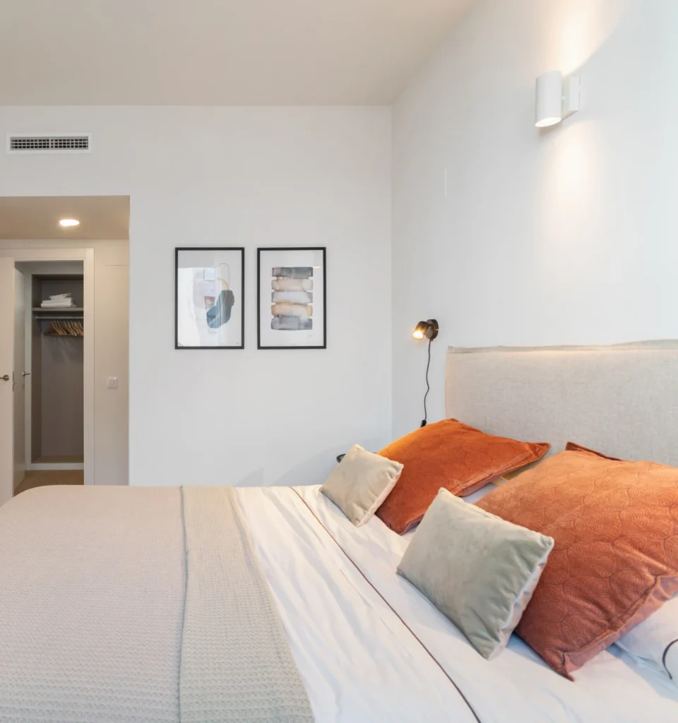 Un dormitorio moderno con una cama de matrimonio, con almohadas naranjas y grisáceas, manta gris, cabecero de color claro, dos obras de arte enmarcadas en una pared blanca y un armario empotrado al fondo.