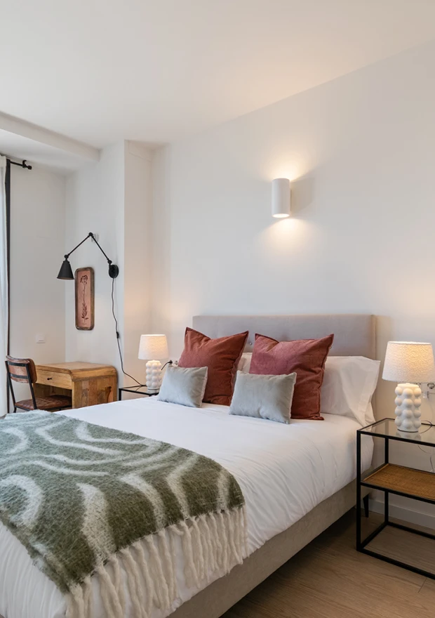 Un dormitorio acogedor con una cama bien hecha con cojines grises y óxido, una manta verde estampada, mesillas de noche con lámparas, un escritorio de madera con una lámpara de pared negra y una decoración minimalista en un espacio luminoso y moderno.
