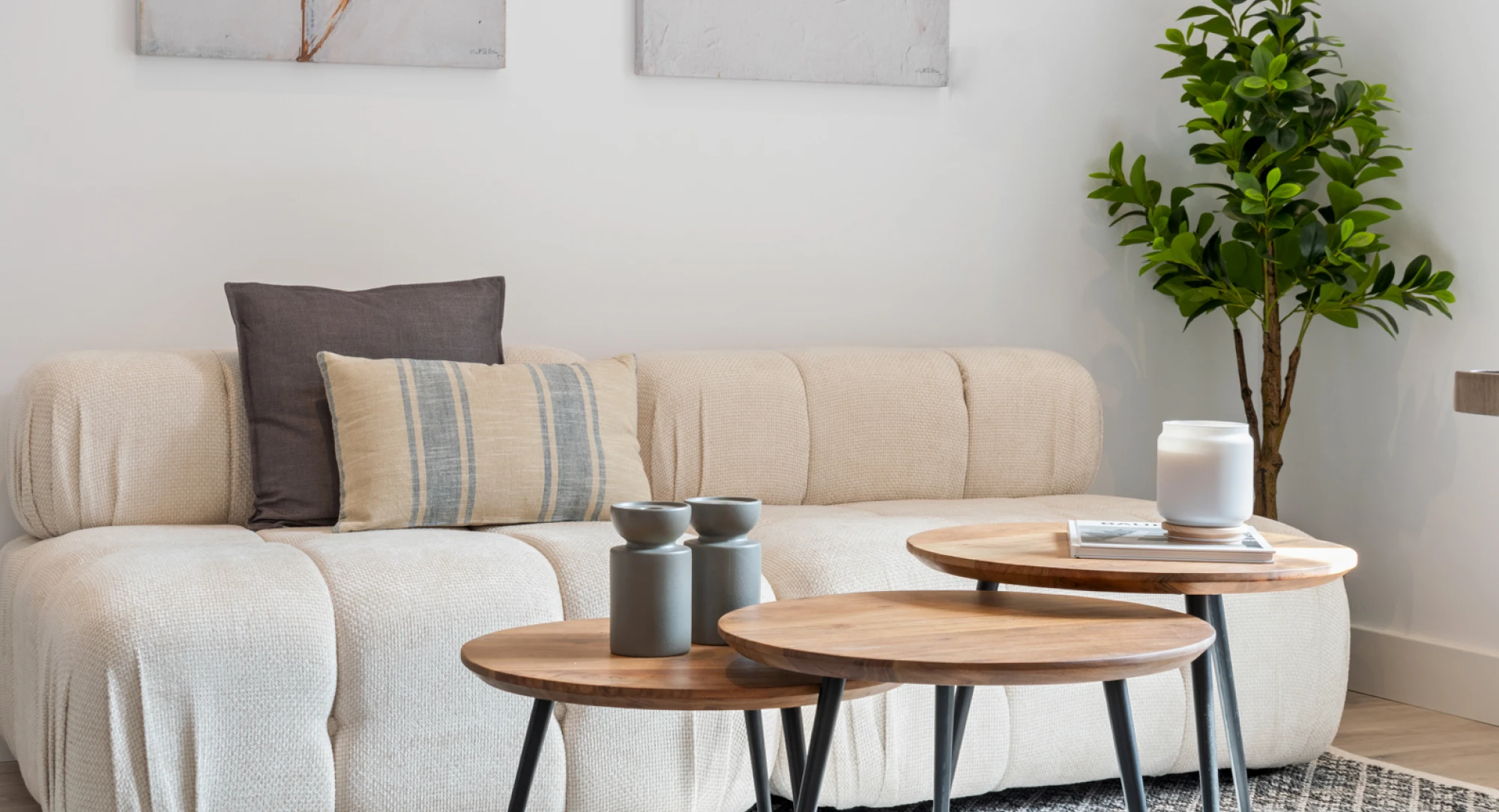 Un salón moderno con un sofá beige, cojines estampados, tres mesas de centro redondas de madera, una planta verde en una maceta y arte mural minimalista al fondo.