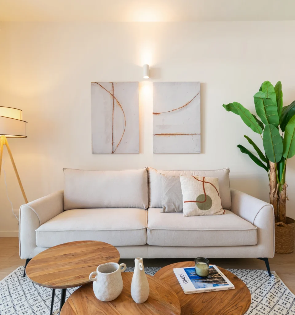 Una moderna sala de estar con un sofá beige, arte en la pared, una lámpara de pie alta con luz cálida, una maceta y mesas de centro redondas de madera anidadas y una alfombra estampada en el suelo de madera.