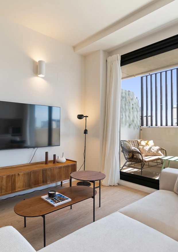 Moderna sala de estar con un televisor montado en la pared, consola de madera, mesas de centro anidadas, sofá luminoso y un balcón con asientos al aire libre, visible a través de puertas correderas de cristal por las que entra luz natural.