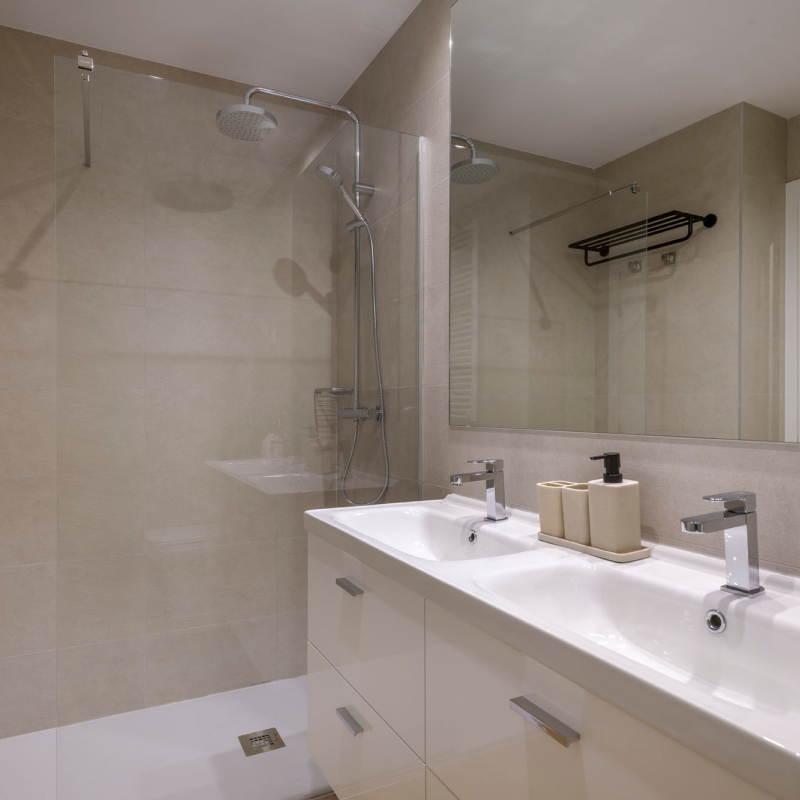 Moderno cuarto de baño con ducha acristalada, cabezal de ducha tipo lluvia, paredes alicatadas en beige, lavabo doble con armarios blancos, espejo grande y dosificadores de jabón beige en la encimera.