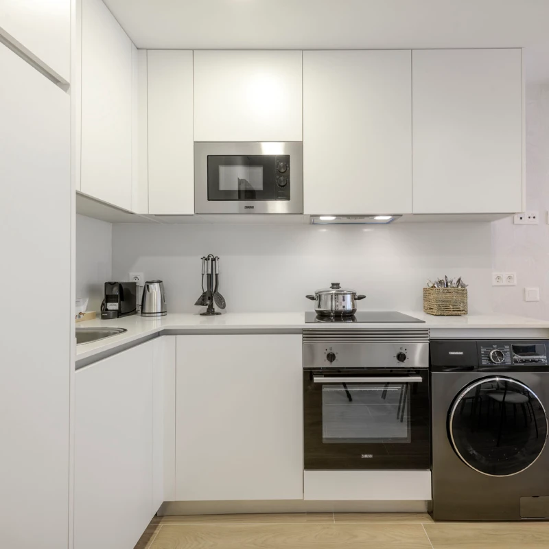 Moderna cocina blanca con electrodomésticos empotrados como horno, microondas y lavadora en color plata. Hay una olla sobre los fogones y pequeños electrodomésticos como un hervidor y una tostadora sobre la encimera.