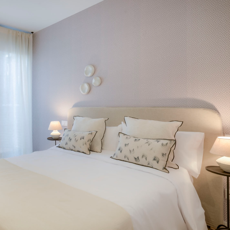 Una cama bien hecha, con sábanas blancas y cojines decorativos, está apoyada en una pared con motivos claros. Sobre el cabecero acolchado cuelgan tres pequeños platos. Dos lámparas sobre mesas auxiliares redondas proporcionan una cálida iluminación. Unas cortinas cubren una ventana.