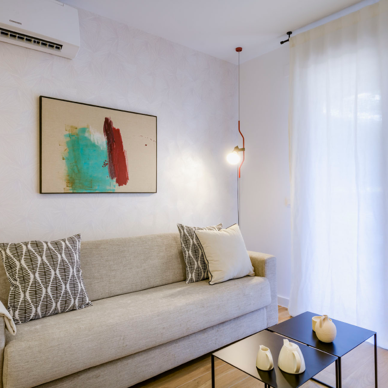 Un salón moderno con un sofá beige, cojines estampados, arte abstracto en la pared, una lámpara colgante, cortinas transparentes y una mesa de centro negra con objetos decorativos. La luz natural llena el espacio.