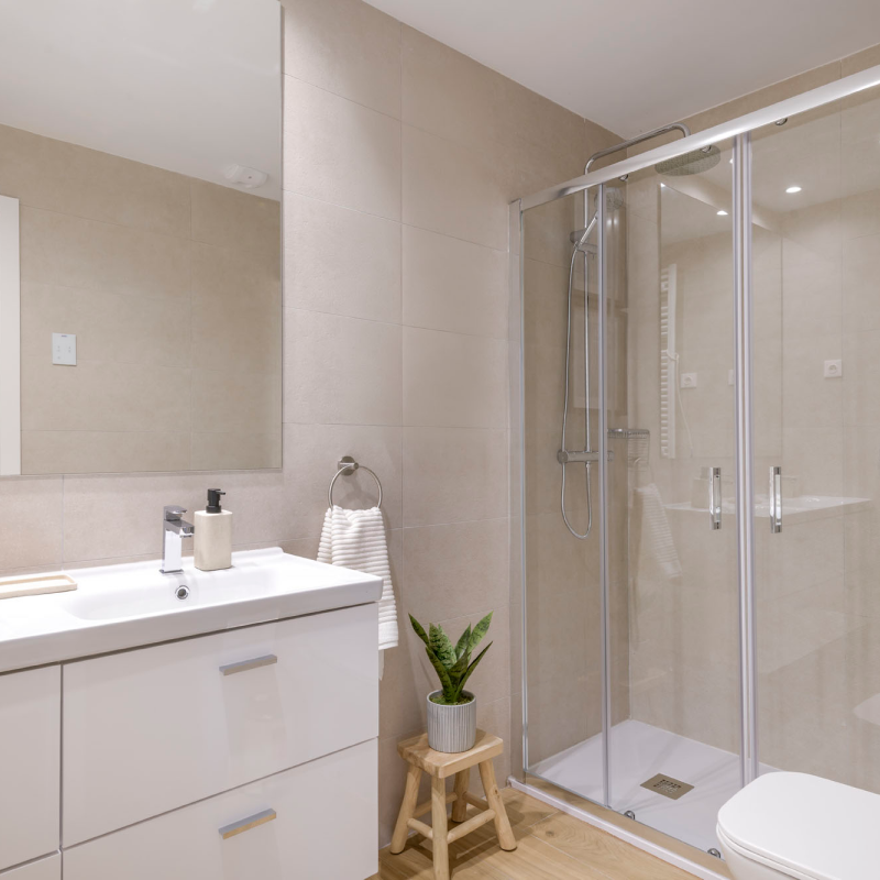 Moderno cuarto de baño con azulejos beige, ducha acristalada, lavabo blanco con pila, dosificador de jabón, toalla en una anilla y una pequeña maceta sobre un taburete de madera. Iluminación clara y decoración minimalista.