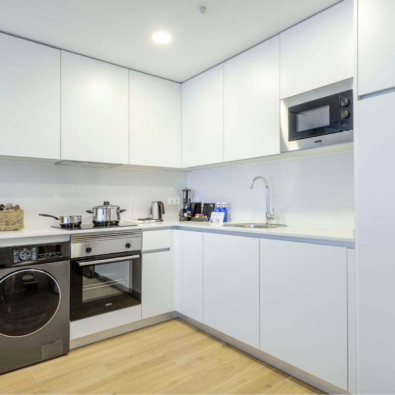 Moderna cocina blanca con electrodomésticos empotrados, incluyendo horno, microondas, lavadora, fregadero y encimera con utensilios de cocina, ollas y pequeños electrodomésticos. Suelo de madera clara e iluminación brillante.