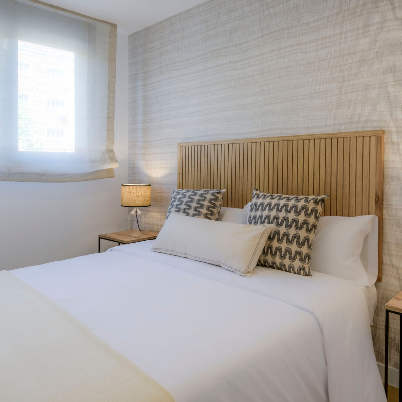Un dormitorio moderno con cabecero de madera, ropa de cama blanca, dos cojines estampados, mesillas de noche con lámparas y una ventana con cortinas transparentes que dejan pasar la luz natural.