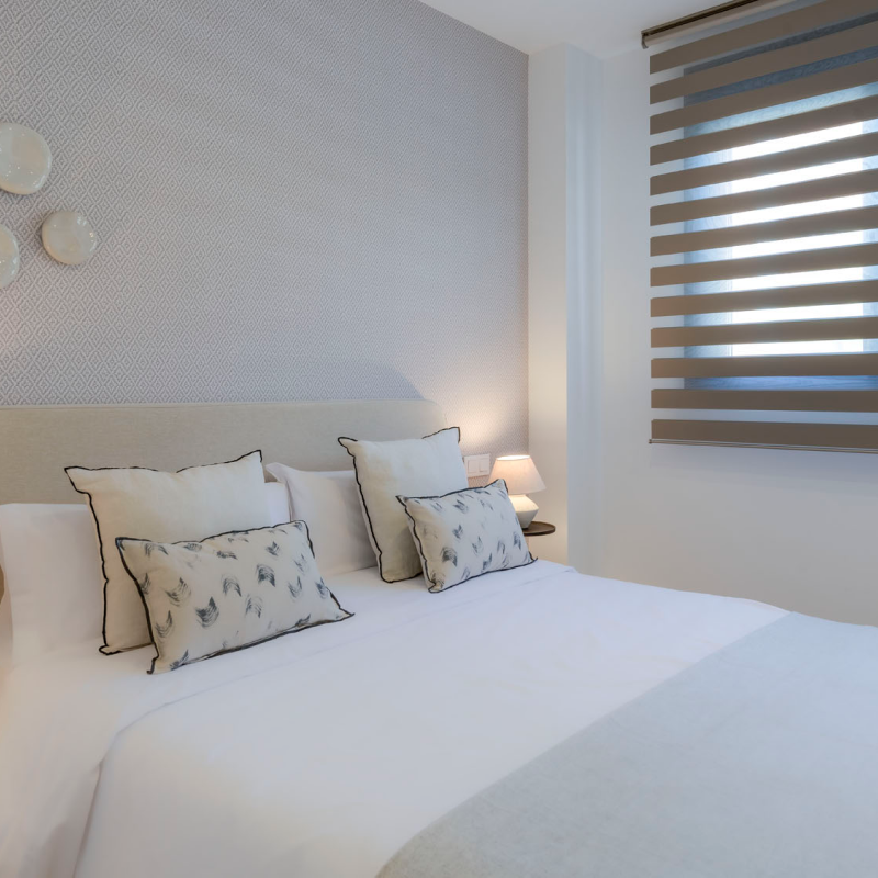 Un dormitorio moderno con una cama bien hecha con almohadas blancas y estampadas, la suave iluminación de una lámpara de mesilla, platos decorativos en la pared y una ventana con persianas de rayas horizontales.