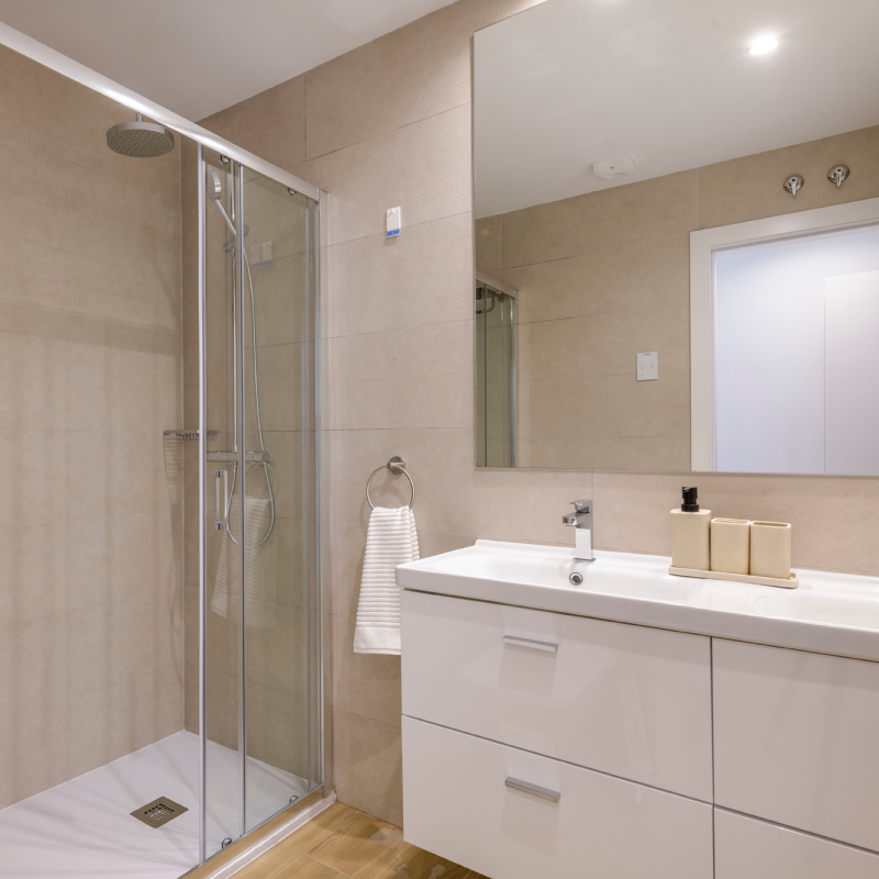 Cuarto de baño con ducha acristalada, paredes alicatadas en beige, un lavabo blanco con cajones, un gran espejo y una toalla colgada de una anilla.