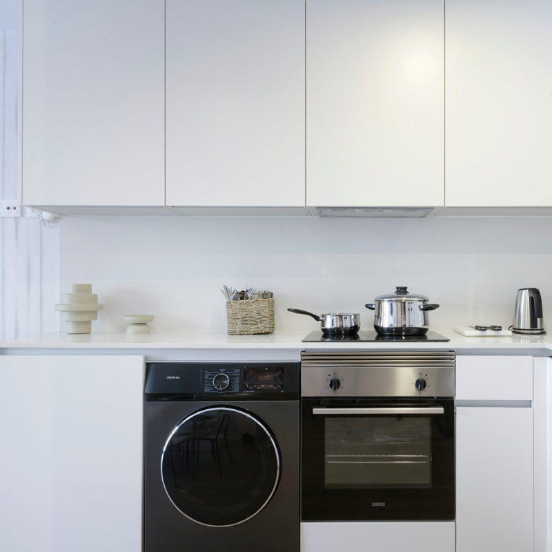 Moderna cocina blanca con electrodomésticos empotrados como horno, microondas y lavadora en color plata. Hay una olla sobre los fogones y pequeños electrodomésticos como un hervidor y una tostadora sobre la encimera.