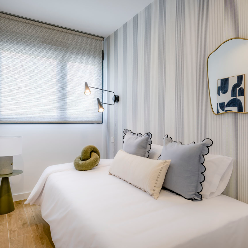 Un dormitorio moderno con una cama individual, ropa de cama blanca, almohadas azules y beige, un cojín de nudo, una lámpara de pared, una mesa auxiliar redonda con una lámpara, un espejo y papel pintado a rayas. La luz inunda la habitación a través de una ventana.