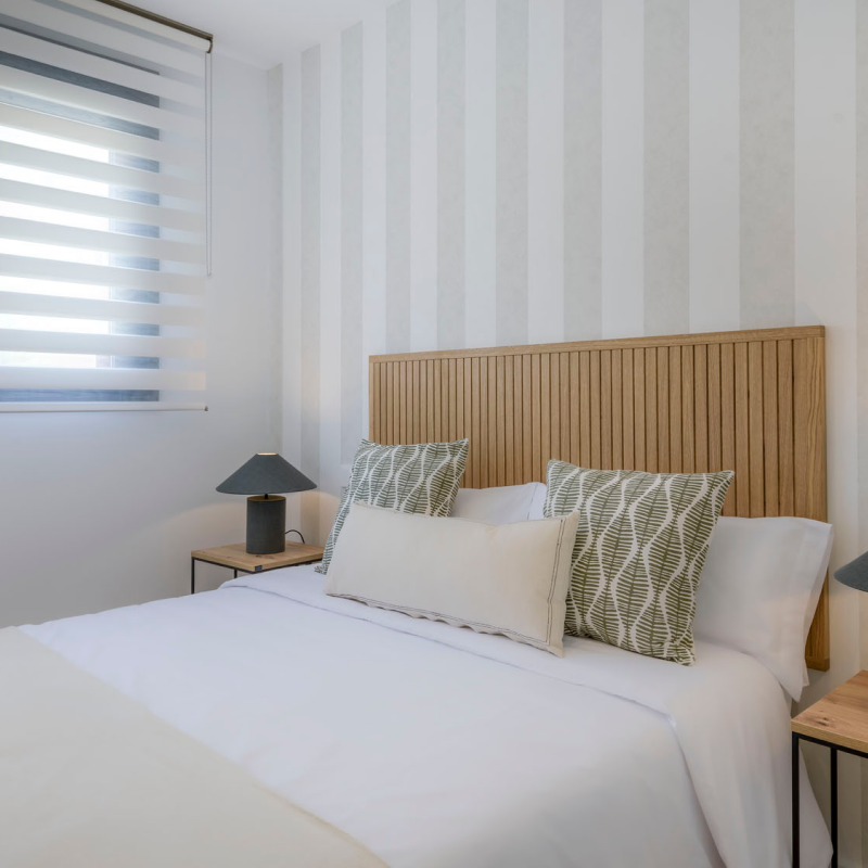 Un dormitorio moderno con cabecero de madera, dos almohadas estampadas y colcha blanca. Hay mesillas de noche a juego con lámparas negras, y una ventana con persianas a rayas deja pasar la luz natural.
