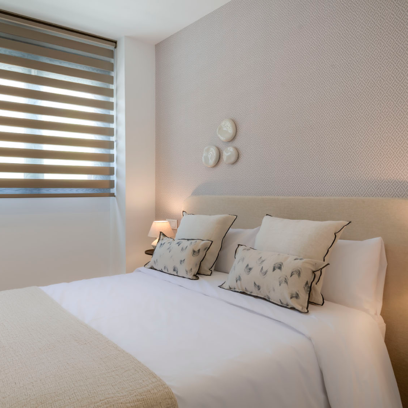 Un dormitorio con una cama individual con cojines estampados, mesillas de noche con lamparas, platos decorativos en la pared y una ventana con persianas.