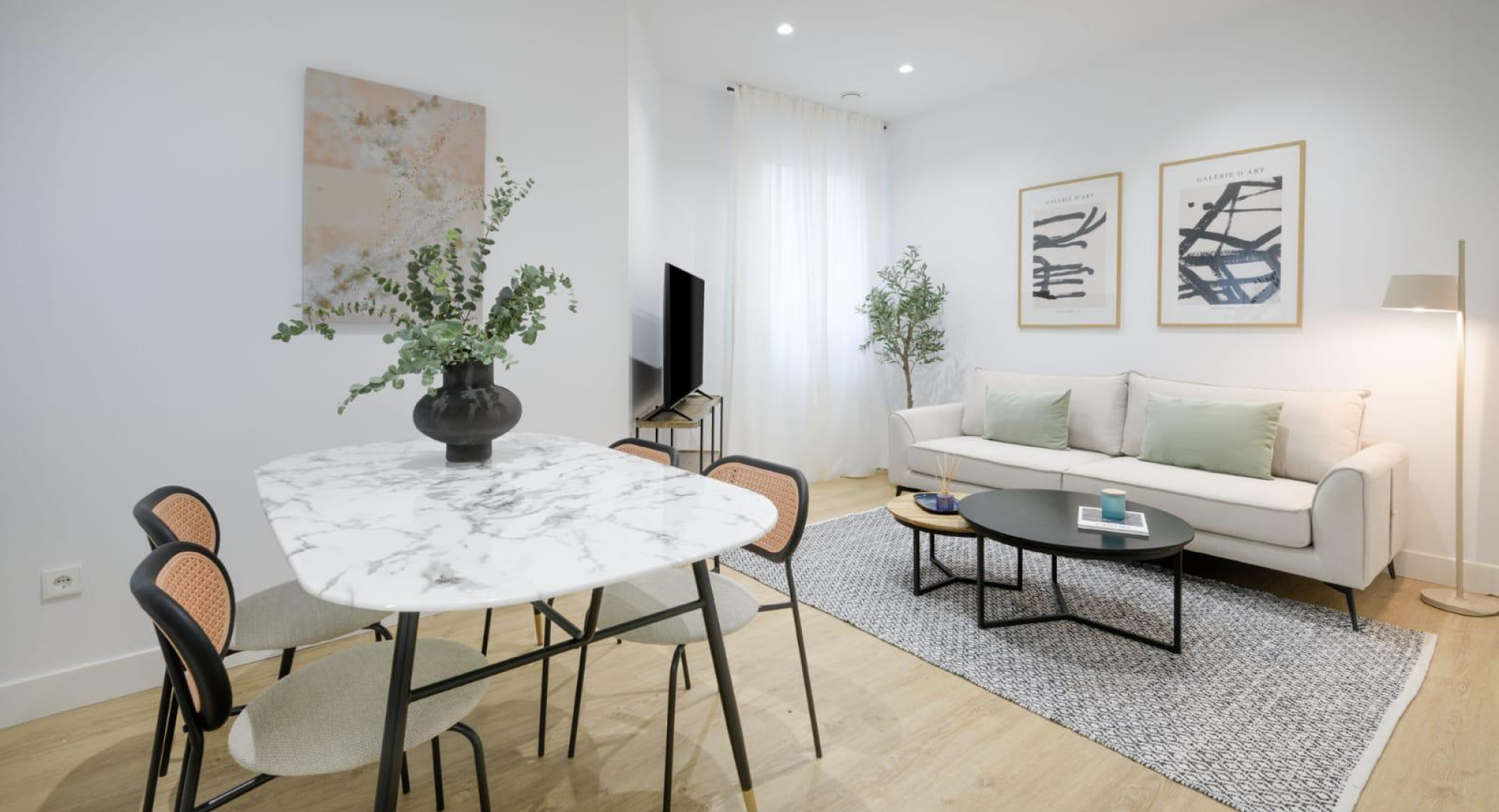 Un salón-comedor moderno y luminoso con una mesa de comedor de mármol, un jarrón negro con plantas, cuatro sillas, un sofá blanco, dos obras de arte abstracto, TV y suelo de madera clara. La luz natural inunda la habitación a través de cortinas transparentes.