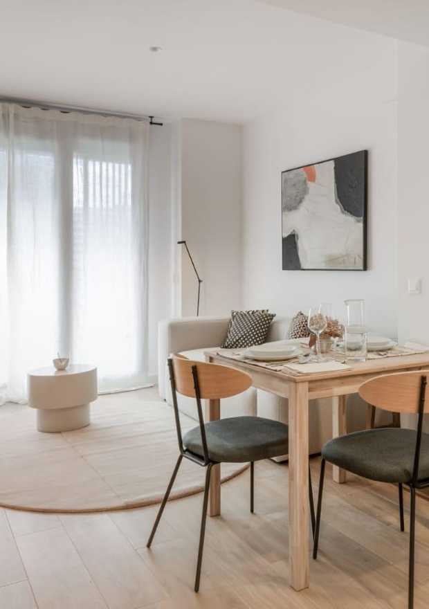 Un comedor moderno y minimalista con una mesa de madera clara para dos personas, sillas negras y de madera, una pequeña mesa de centro redonda y un acogedor sofá cerca de cortinas transparentes y arte abstracto en la pared.