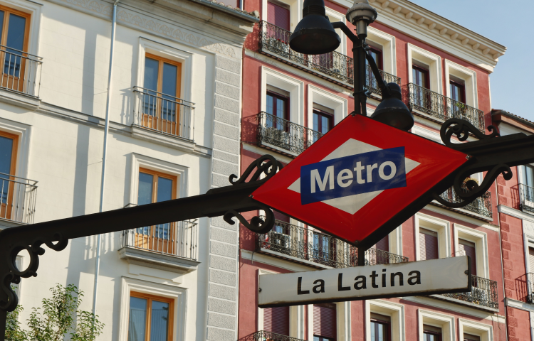 Un cartel del Metro de Madrid cuelga sobre una calle, con La Latina debajo. Al fondo se ven edificios con fachadas coloridas.