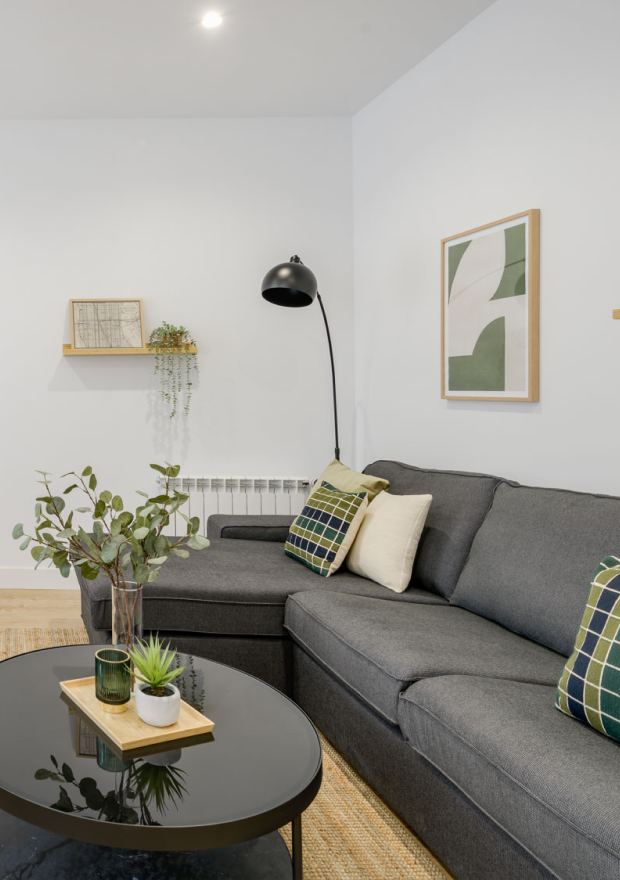 Salón moderno con un sofá seccional gris oscuro, cojines verdes y crema, una mesa de centro negra redonda con plantas y velas, arte minimalista en la pared, una pequeña estantería con decoración y una lámpara de pie negra contra una pared blanca.