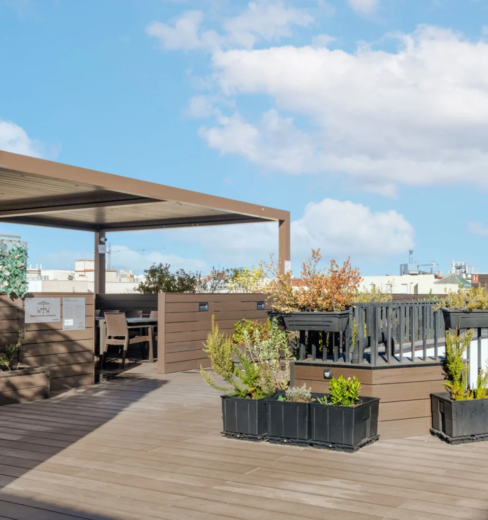 Una moderna terraza en la azotea presenta suelo de madera, jardineras negras con plantas verdes, una pérgola que cubre una zona de asientos y una barandilla para el jacuzzi, todo ello bajo un cielo azul brillante con nubes dispersas.
